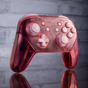 May include: A translucent pink Nintendo Switch controller with white buttons. The controller has a textured surface and is designed for gaming.