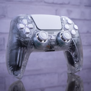 Puede incluir: Un mando de juego PlayStation 5 DualSense transparente blanco con un diseño translúcido. El mando tiene un panel táctil blanco y botones blancos.