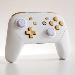 Puede incluir: Un mando de Nintendo Switch blanco con botones dorados y palancas analógicas moradas.