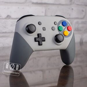 May include: A grey and white Nintendo Switch controller with a custom faceplate featuring the classic Super Nintendo Entertainment System (SNES) button layout. The buttons are coloured red, blue, yellow, and green. The controller is on a wooden surface.