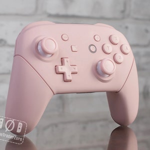 May include: A pink Nintendo Switch Joy-Con controller with white buttons. The controller is on a reflective surface.