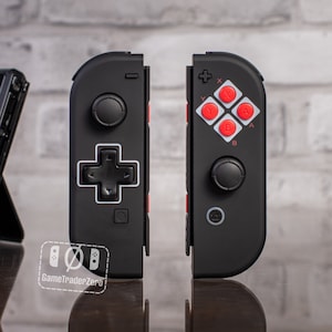 May include: A black Nintendo Switch controller with a red and white NES-style button layout. The controller has a D-pad, two analog sticks, and four face buttons. The text "GameTraderZero" is visible on the controller.