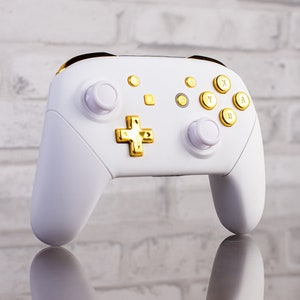 Könnte beinhalten: Ein weißer Nintendo Switch-Controller mit goldenen Knöpfen und Akzenten. Der Controller hat einen weißen Hintergrund mit goldenen Knöpfen und Akzenten. Der Controller befindet sich auf einem weißen Ziegelhintergrund.
