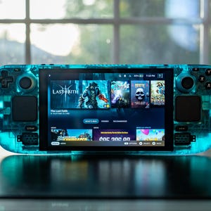 May include: A transparent teal handheld gaming console displaying a game library on its screen. The console has buttons, joysticks, and a screen showing game titles. The device is on a dark surface with a blurred background.