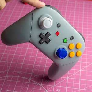 Puede incluir: Un mando de videojuegos gris con un botón blanco, un botón azul y cuatro botones amarillos. El mando tiene un pad direccional y un botón rojo.