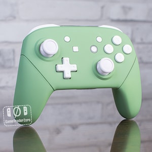 May include: A green and white Nintendo Switch controller with white buttons and a white plus sign button. The controller is on a wooden surface.