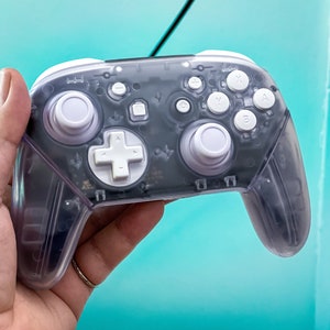 May include: A clear, translucent, white Nintendo Switch controller with black buttons and a white plus sign button. The controller has a textured surface and is held in a hand.