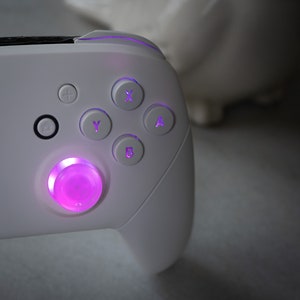 May include: A white gaming controller with a purple glowing button in the center. The controller has buttons labeled A, B, X, Y, and a directional pad.