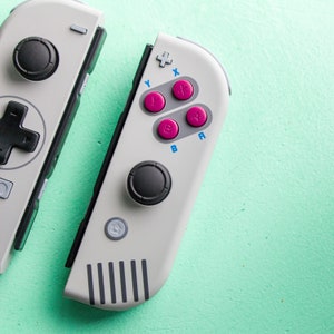 Może przedstawiać: Biało-szary kontroler Nintendo Switch w retro stylu. Kontroler posiada czarny joystick, krzyżak i cztery r&oacute;żowe przyciski oznaczone literami "Y", "X", "A" i "B".