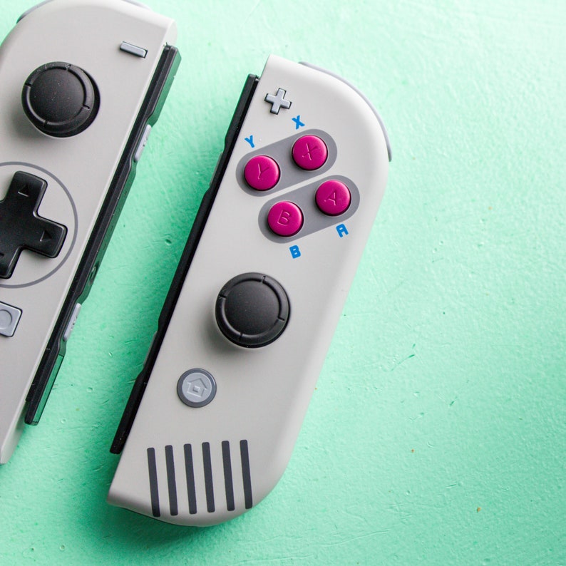 Może przedstawiać: Biało-szary kontroler Nintendo Switch w retro stylu. Kontroler posiada czarny joystick, krzyżak i cztery r&oacute;żowe przyciski oznaczone literami "Y", "X", "A" i "B".