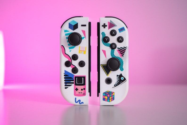 May include: A pair of white Nintendo Switch Joy-Con controllers with a retro 80s themed design. The controllers feature a variety of colorful geometric shapes, including a Rubik's Cube, a sneaker, and a pixelated video game character. The controllers are separated, showing the left and right sides.
