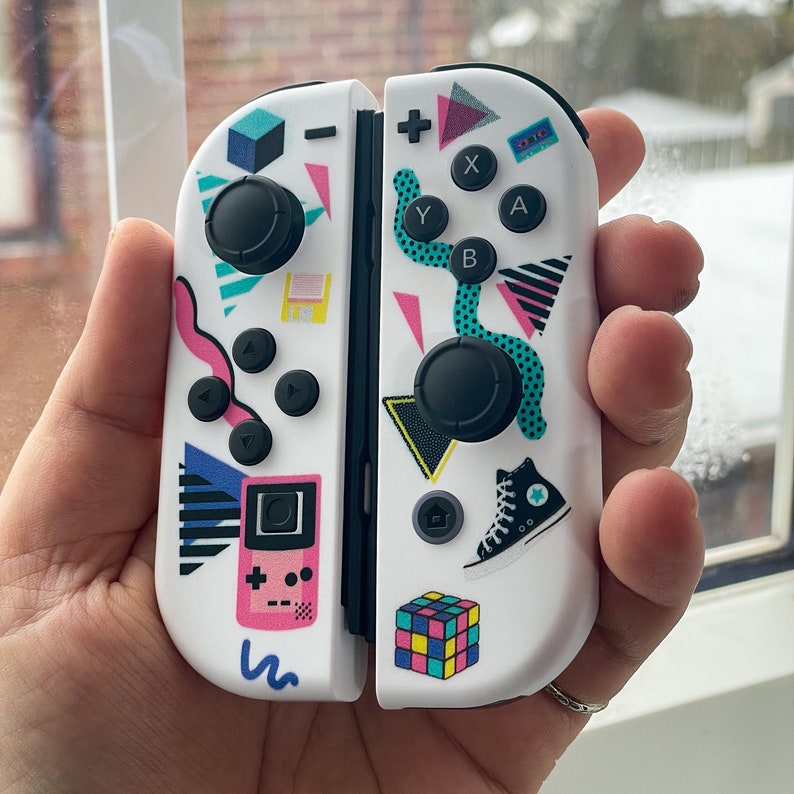 May include: White Nintendo Switch Joy-Con controllers with a retro 80s themed design. The design features a pink and blue color scheme with geometric shapes, a cassette tape, a game cartridge, a Game Boy, and a Rubik's Cube.