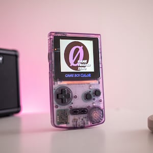May include: A purple Game Boy Color handheld video game console with a black and white screen displaying the logo "Game Trader Zero" with a stylized letter "O".