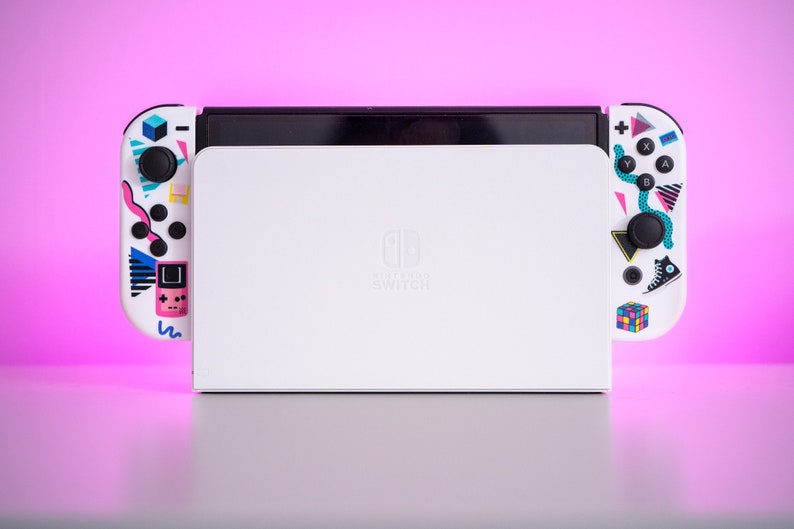 May include: A white Nintendo Switch console with a retro-themed design. The console has a black screen and two white Joy-Con controllers with colorful 80s-inspired graphics. The text "Nintendo Switch" is printed on the console.