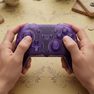 Puede incluir: Un mando de videojuegos morado transparente con varios botones y un panel direccional. El mando est&aacute; sujeto por dos manos, mostrando su dise&ntilde;o. Los botones tambi&eacute;n son morados.