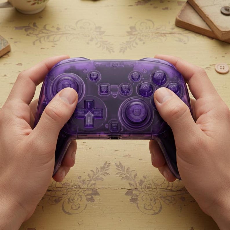Puede incluir: Un mando de videojuegos morado transparente con varios botones y un panel direccional. El mando est&aacute; sujeto por dos manos, mostrando su dise&ntilde;o. Los botones tambi&eacute;n son morados.