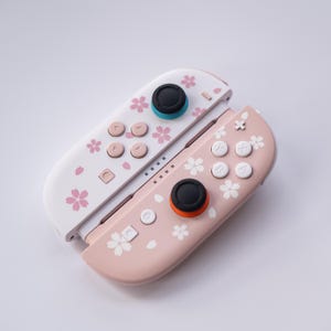 May include: Two pastel-colored game controllers. One is white, the other is pink. Both controllers feature a floral cherry blossom design. The buttons are white, pink, and black. The controllers are designed for gaming.