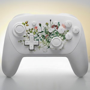 May include: A white video game controller with a floral design. The controller features a botanical pattern of green leaves, pink and red flowers, and small white buttons. The controller has two joysticks, a directional pad, and several buttons labeled X, Y, A, and B.