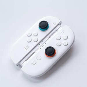 May include: White gaming controllers with black and colored accents. The controllers have multiple buttons and directional pads. The joysticks have black tops with blue and orange rings. The controllers are designed for a gaming console.