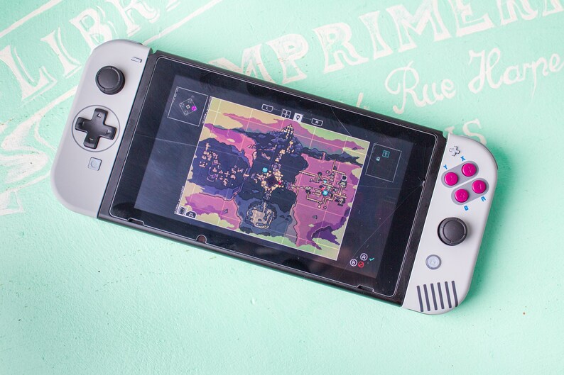 Może przedstawiać: Szara konsola Nintendo Switch z czarnym ekranem wyświetlającym mapę. Konsola ma spersonalizowaną płytkę czołową w kolorze szarym i czarnym oraz zestaw r&oacute;żowych i niebieskich przycisk&oacute;w.
