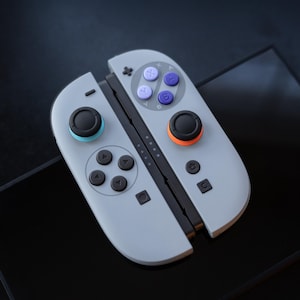 May include: A pair of gray game controllers with black joysticks and colored buttons. The left joystick has a blue ring, and the right joystick has an orange ring. The controllers are open, revealing the connection points.