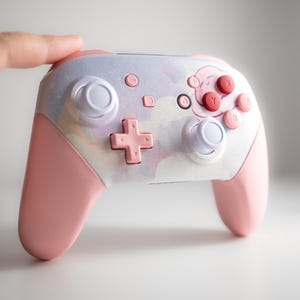 May include: A pink and white Nintendo Switch controller with a custom design. The controller has a pink and white color scheme with a white and pink button layout. The controller has a white and pink button layout.