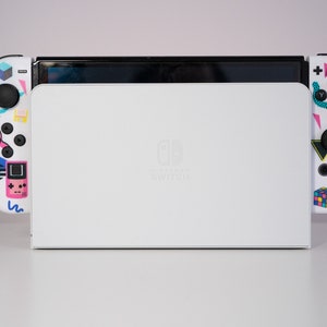 May include: A white Nintendo Switch console with colorful retro-themed designs on the controllers. The console is closed and the Nintendo Switch logo is visible on the front.