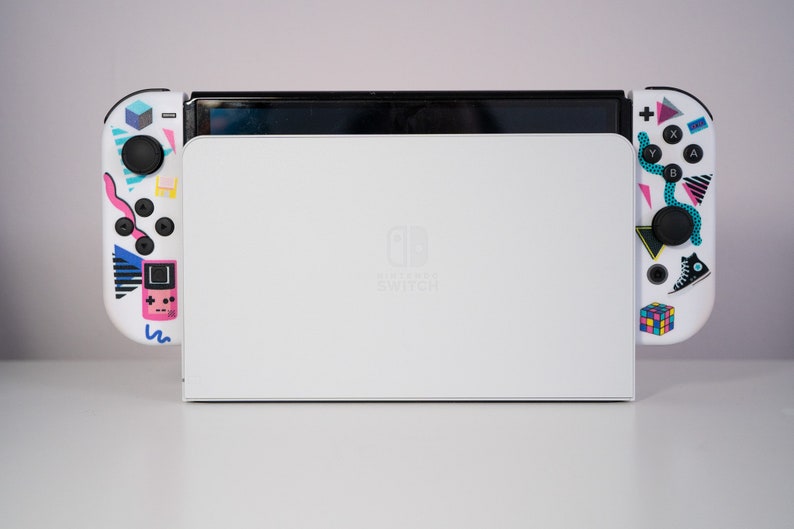May include: A white Nintendo Switch console with colorful retro-themed designs on the controllers. The console is closed and the Nintendo Switch logo is visible on the front.