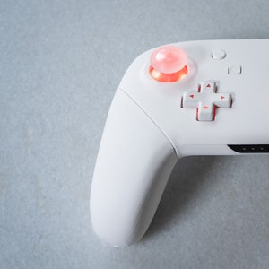 May include: A white wireless gaming controller with a red button and a directional pad. The controller has a smooth, rounded design.