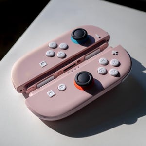Könnte beinhalten: Zwei rosa Gamecontroller mit weißen Knöpfen und schwarzen Joysticks. Ein Joystick hat einen blauen Ring, der andere einen orangefarbenen Ring. Die Controller liegen auf einer weißen Oberfläche.