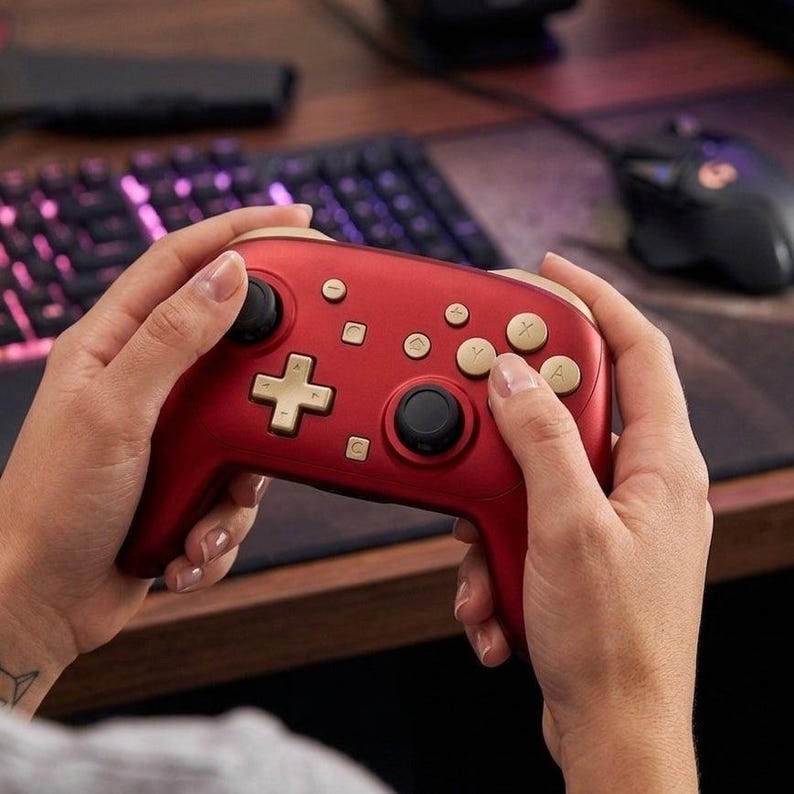 May include: A matte red wireless video game controller featuring gold-colored buttons, a gold directional pad, and two black analog sticks. The controller includes a central home button, a capture button, and four primary action buttons labeled A, B, X, and Y.