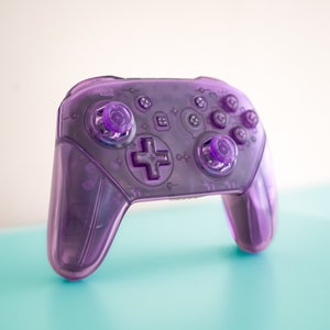 Puede incluir: Un mando de videojuegos translúcido de color morado con un botón en forma de cruz y varios botones redondos.