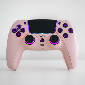 May include: A pink PlayStation 5 DualSense controller with purple glowing buttons. The controller has a black touchpad and a PlayStation logo in the centre.