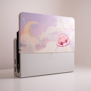 May include: A white Nintendo Switch console with a pink and purple custom skin featuring a cartoon character sleeping on a cloud. The skin has a watercolor effect and depicts a crescent moon and stars.