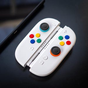 Könnte beinhalten: Weiße Gaming-Controller mit farbigen Knöpfen und schwarzen Joysticks. Die Controller haben rote, gelbe, grüne und blaue Knöpfe. Die Joysticks haben blaue und orangefarbene Akzente. Im Hintergrund befindet sich ein schwarzes Smartphone.