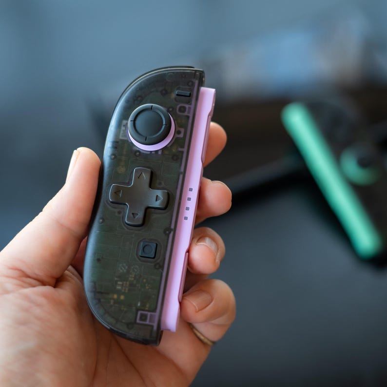 Puede incluir: Un mando de videojuegos negro transparente y morado. El mando tiene un panel direccional, botones y un joystick negros. El lado derecho es de color morado claro s&oacute;lido. El mando se sostiene en la mano de una persona.