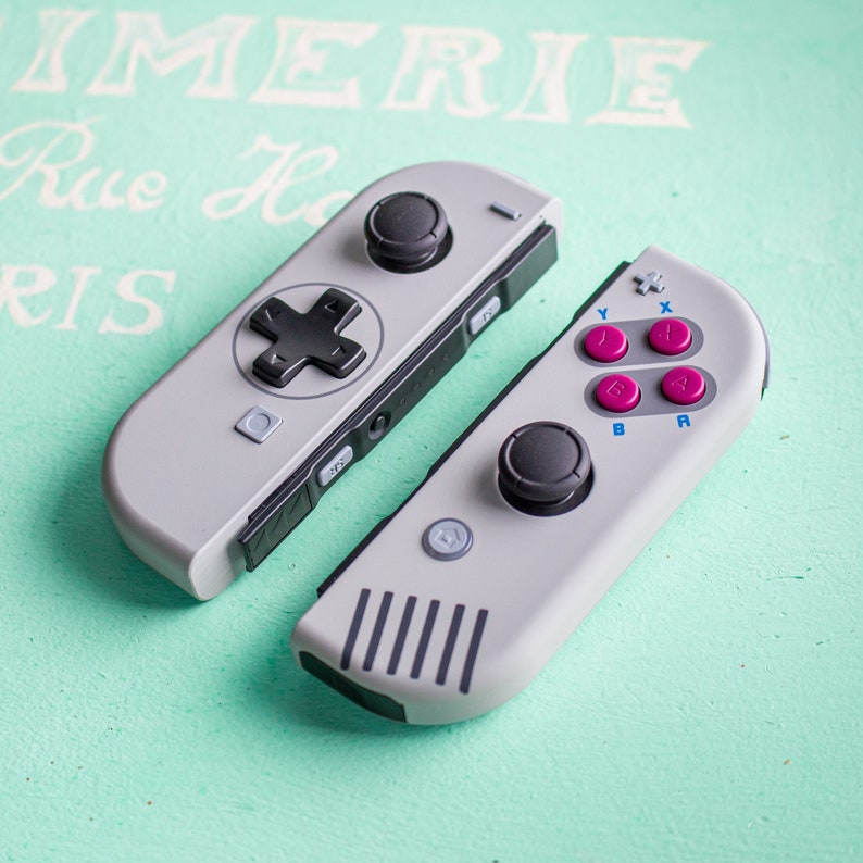 Może przedstawiać: Para szaro-czarnych kontroler&oacute;w Nintendo Switch Joy-Con w retro stylu. Lewy kontroler ma czarny krzyż kierunkowy, a prawy kontroler ma czarny analogowy joystick i cztery kolorowe przyciski oznaczone literami Y, X, A i B.