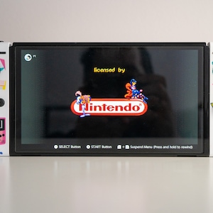 May include: A white Nintendo Switch console with a retro-themed design. The console is displaying a screen with the text "licensed by Nintendo".