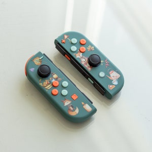 May include: Two green Nintendo Switch Joy-Con controllers with custom designs. One controller features a fox, teapot, and other autumnal imagery. The other controller features a fox, a candle, and other autumnal imagery.