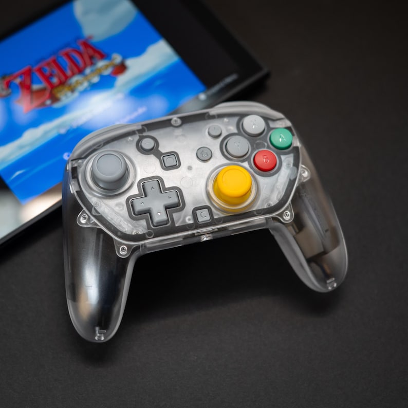 May include: A transparent gaming controller with gray, yellow, red, and green buttons. The controller is positioned in front of a screen displaying a video game. The controller has a clear plastic shell.