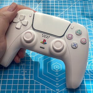 May include: A white PlayStation 5 controller featuring the PlayStation logo in colour. The controller has a touchpad, directional buttons, and analogue sticks. The buttons are coloured green, pink, blue, and purple.