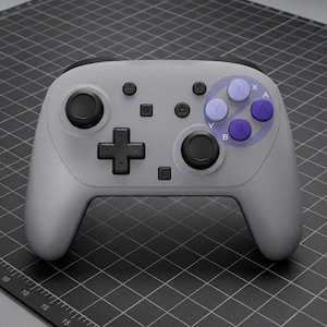 May include: A gray wireless game controller with black buttons and a directional pad. The controller has purple and light purple buttons on the right side. The controller is on a black grid surface.