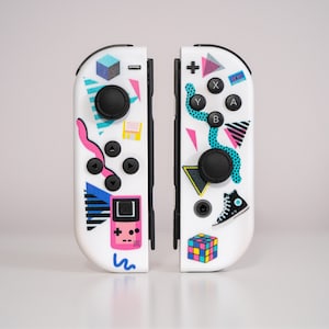 May include: A pair of white Nintendo Switch Joy-Con controllers with colorful 80s-themed designs. One controller features a pink and blue geometric pattern, a game cartridge, and a pink and blue snake. The other controller features a blue and green geometric pattern, a black and white sneaker, and a Rubik's Cube.