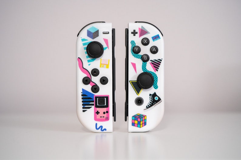 May include: A pair of white Nintendo Switch Joy-Con controllers with colorful 80s-themed designs. One controller features a pink and blue geometric pattern, a game cartridge, and a pink and blue snake. The other controller features a blue and green geometric pattern, a black and white sneaker, and a Rubik's Cube.