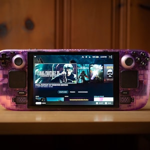 Könnte beinhalten: Handheld-Spielkonsole mit transparentem lila Gehäuse, die ein Spielmenü auf ihrem Bildschirm anzeigt. Die Konsole hat schwarze Steuertasten und einen Bildschirm mit Spielen wie "Final Fantasy XV Windows Edition". Sie liegt auf einer Holzoberfläche.