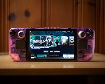 May include: A handheld gaming console with a transparent purple shell, displaying a game menu on its screen. The console has black control buttons and a screen showing game titles like "Final Fantasy XV Windows Edition". It rests on a wooden surface.