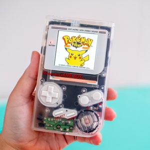 Könnte beinhalten: Eine durchsichtige, transluzente Game Boy Color Handheld-Videospielkonsole mit einem schwarzen Bildschirm, auf dem die Pokemon Yellow-Spielkassette angezeigt wird. Der Bildschirm zeigt den Text "Pokemon Yellow Version" und einen gelben Pikachu-Charakter. Die Konsole hat einen weißen Knopf und einen roten Knopf.