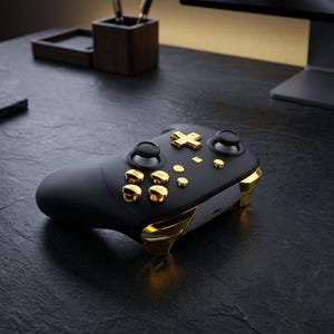 May include: A black and gold wireless game controller. The controller features gold accents on the buttons and edges, with a matte black finish. The controller is on a dark surface.