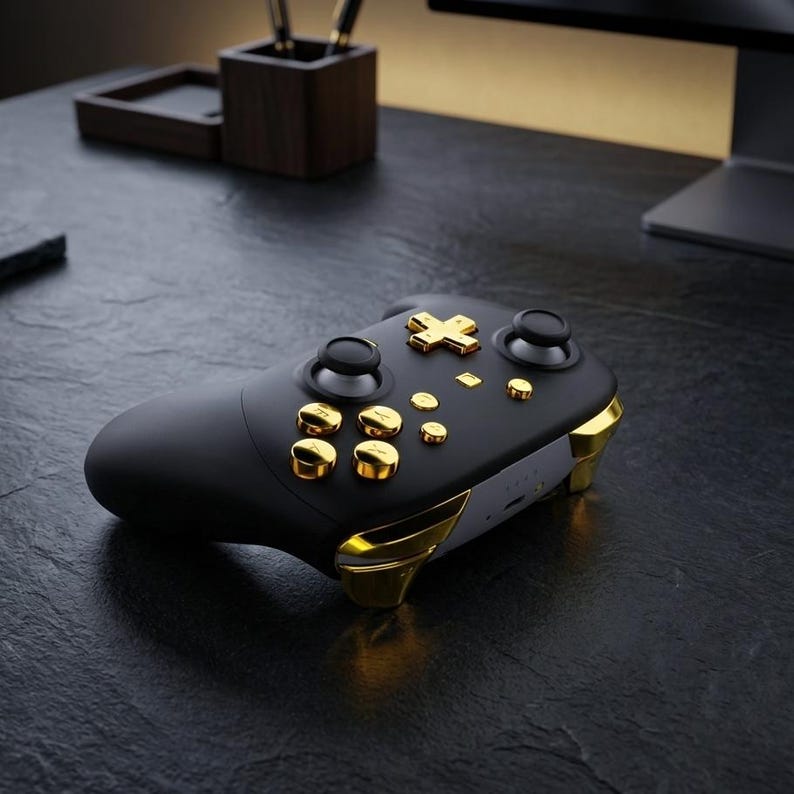 May include: A black and gold wireless game controller. The controller features gold accents on the buttons and edges, with a matte black finish. The controller is on a dark surface.