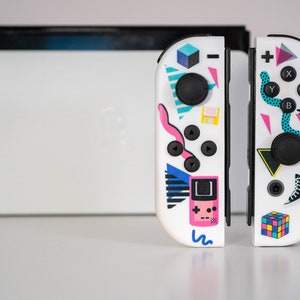 May include: A white Nintendo Switch controller with a retro 80s and 90s themed design. The controller features a variety of colorful geometric shapes, including a pink Game Boy, a blue and white sneaker, and a rainbow Rubik's Cube.